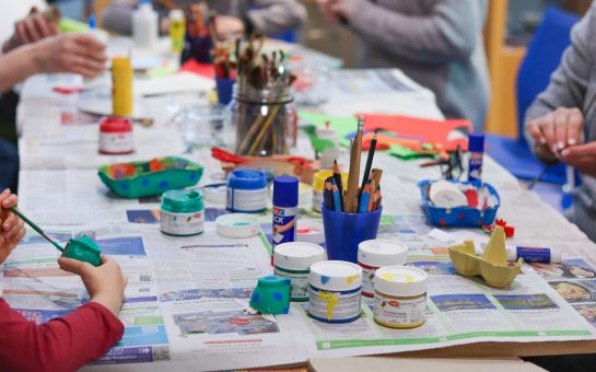 Zus sehen sind bastelnde Kinder, die an einem langen Tisch sitzen. Außerdem Becher mit Buntstiften, Klebestifte und weitere Bastelmaterialien.