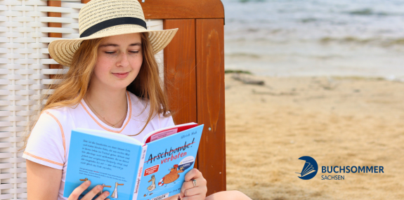 Zu sehen ist ein Mädchen am Strand, welches in einem Buch liest.