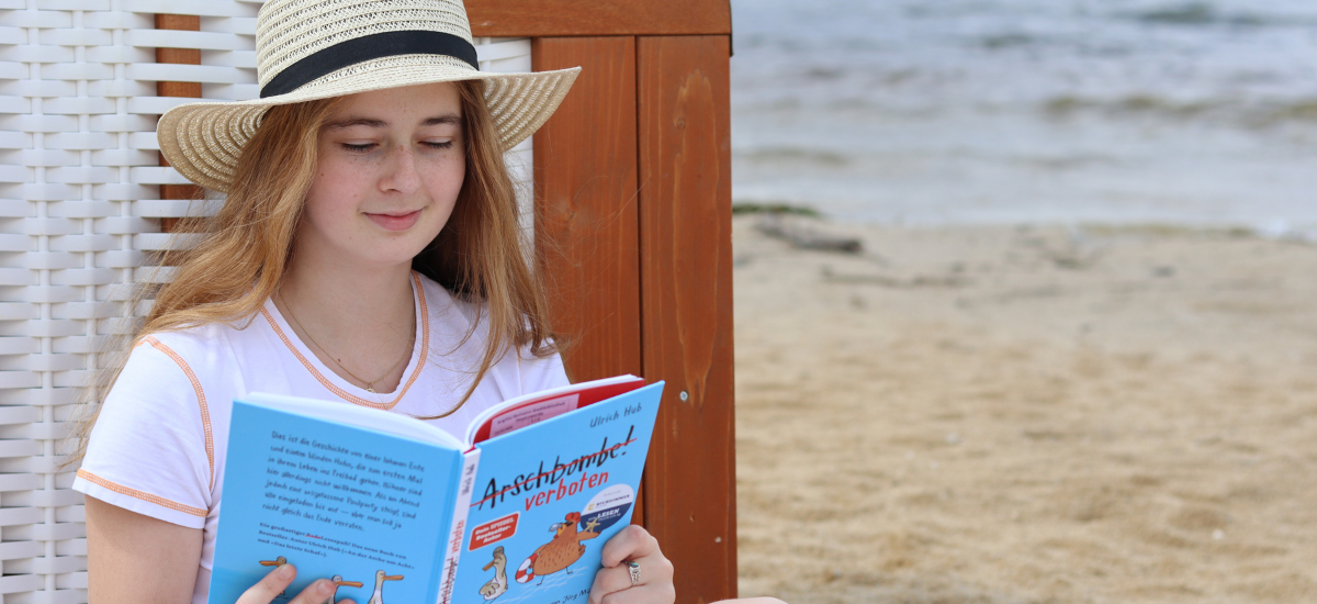 Junge Frau liest am Strand an einem Strandkorb gelehnt in einem Buch.