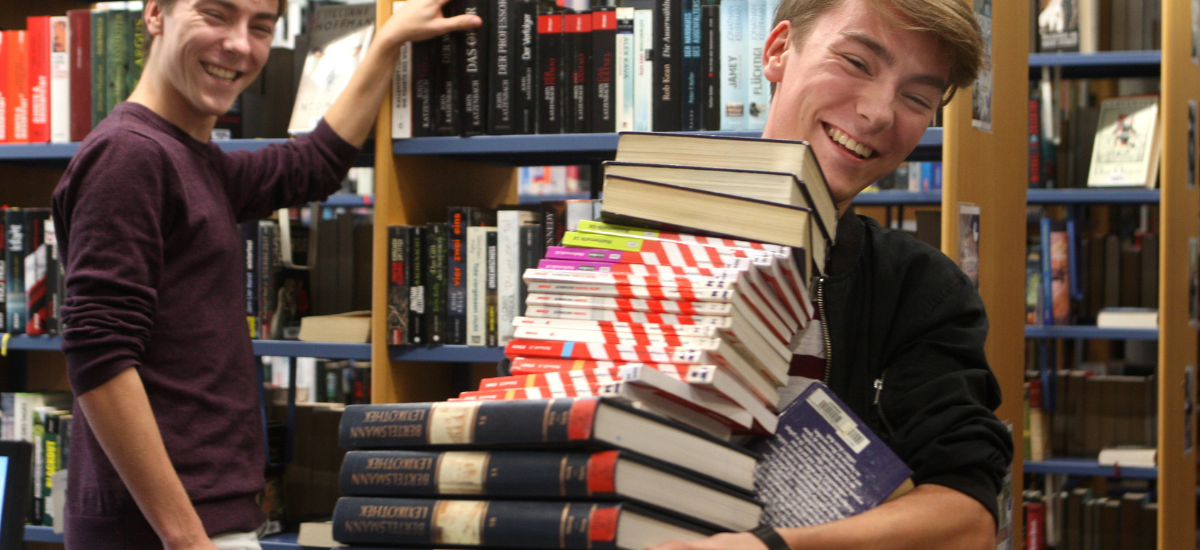 Zwei junge Männer tragen in der Bibliothek einen Stapel von Enzyklopädien